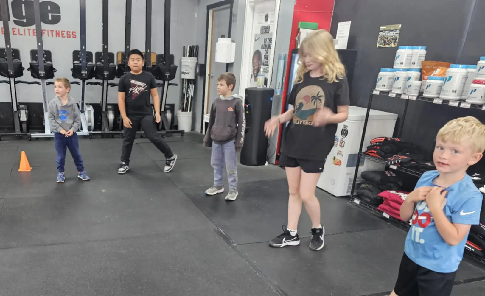 Happy children of various ages participating in a CrossFit Kids class, doing fun bodyweight exercises like bear crawls and jumping, with big smiles and high energy in a colorful, safe gym environment