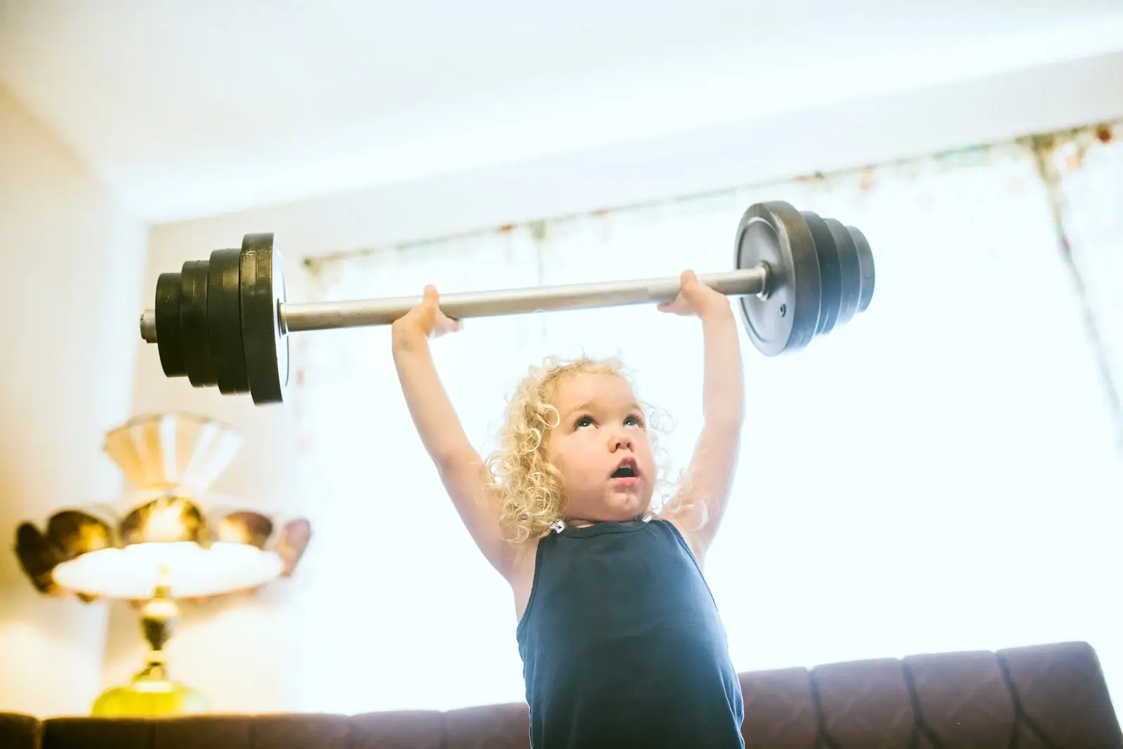 Professional CrossFit Kids coach demonstrating proper squat form to a small group of attentive children (ages 8-12). Coach is kneeling at kid's eye level, showing encouraging body language. Children look engaged and happy in a bright, safe gym environment with age-appropriate equipment visible in background.